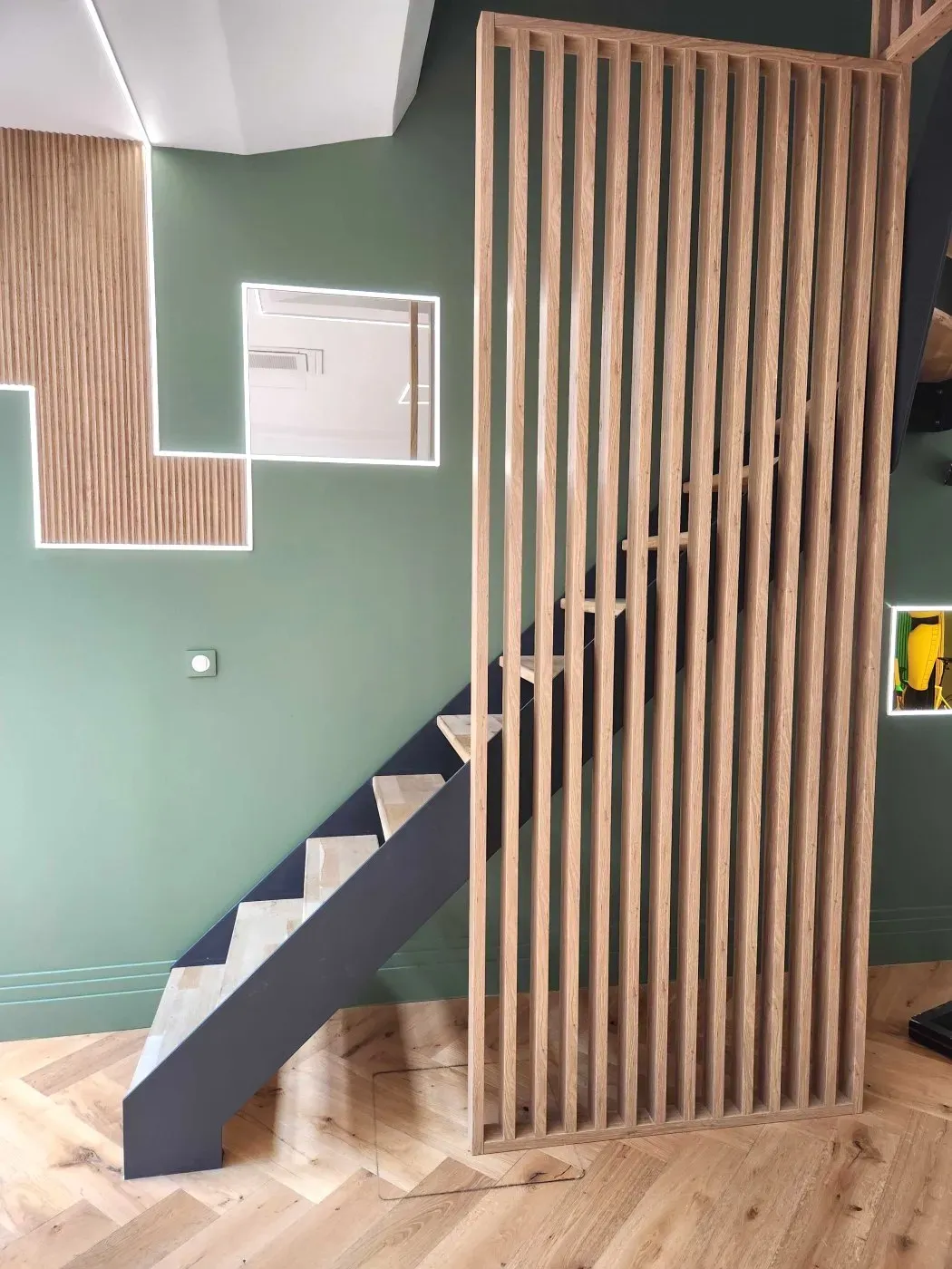 Escalier bordé d'un claustra en tasseaux de bois, devant un mur vert